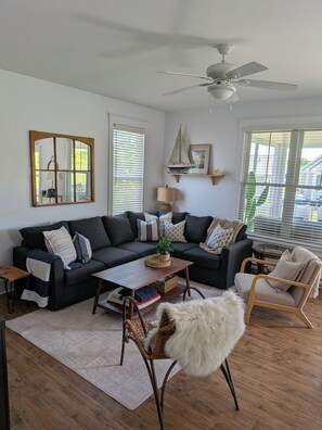 Un salon avec un canapé de couleur foncée, une table basse en bois, un fauteuil blanc à poils longs, un ventilateur de plafond et une grande fenêtre avec des stores.
