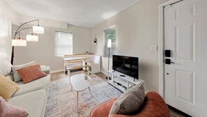 A living room with a sofa, a coffee table, a TV, and a door. - Airport, Stadiums & City Life in ATL! (Atlanta)