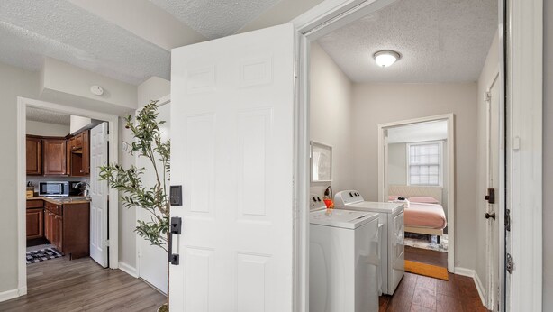 A hallway with a white door leading to a laundry room with a washing machine and dryer, a kitchen area with wooden cabinets, and a small plant. - Airport, Stadiums & City Life in ATL! (Atlanta)
