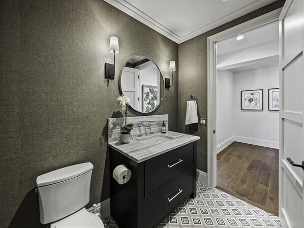 A modern bathroom with a marble sink, black vanity, and a toilet. There is a mirror with a round frame and wall-mounted lights. The floor has a geometric pattern.