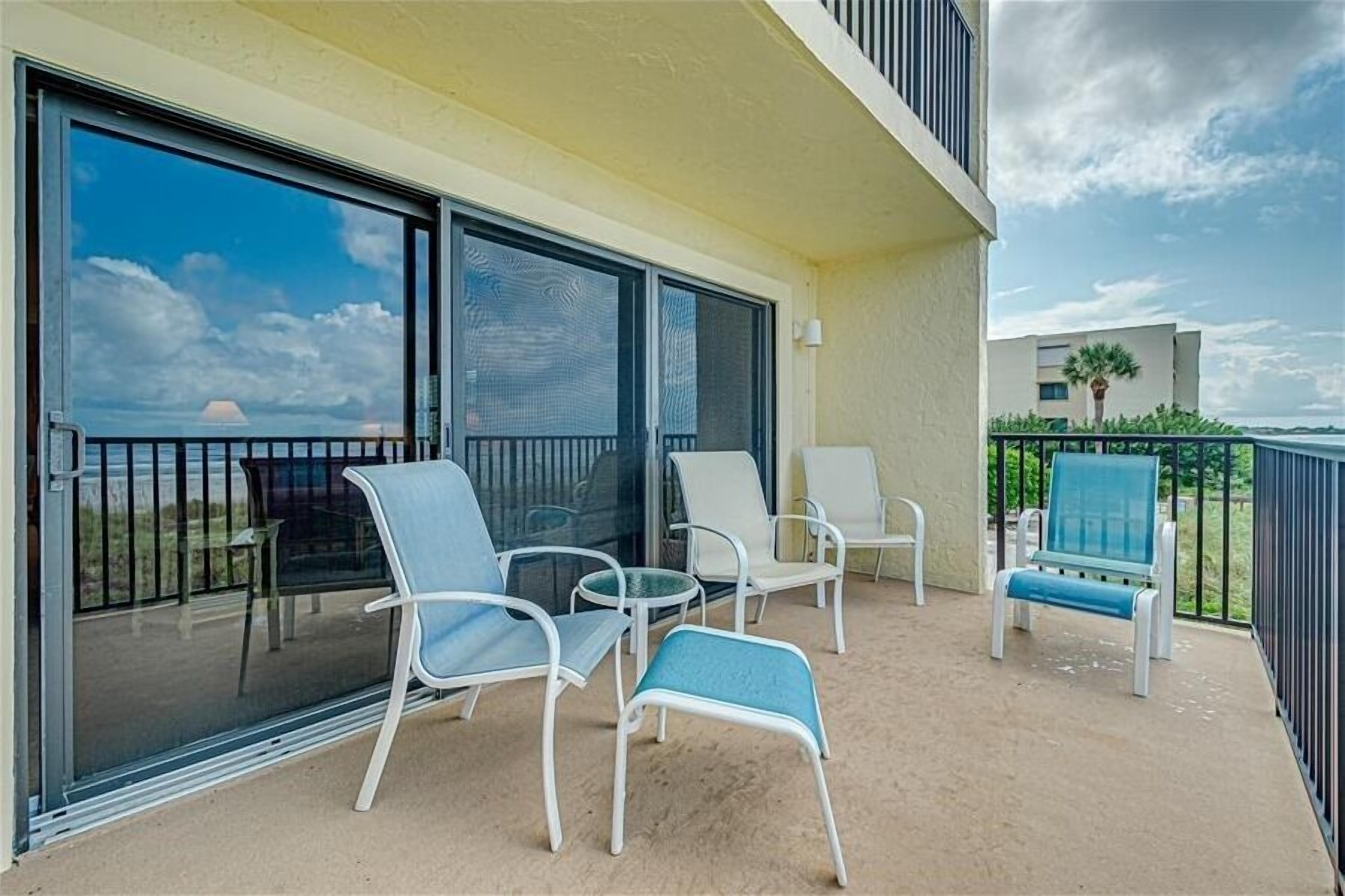 Un balcon avec des chaises en métal blanc et un fauteuil bleu rembourré, une porte vitrée donnant sur une piscine, et une vue sur l’océan et un palmier.