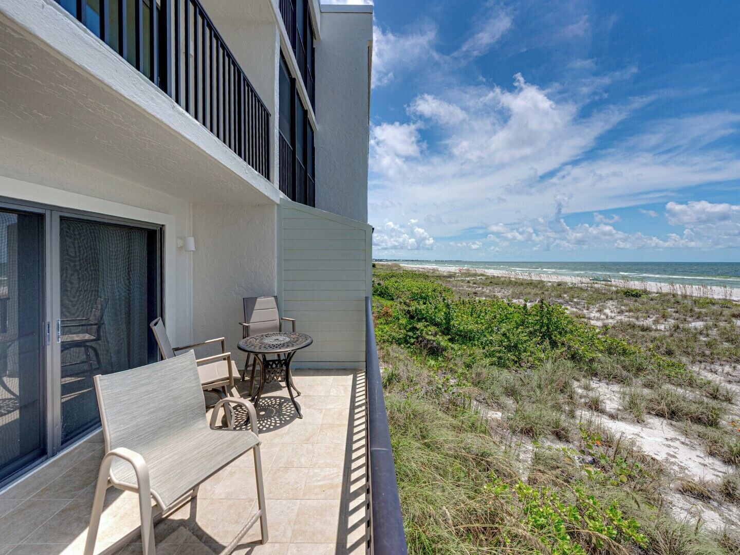 A balcony with a table and chairs, overlooking a beach with sand dunes and the ocean.