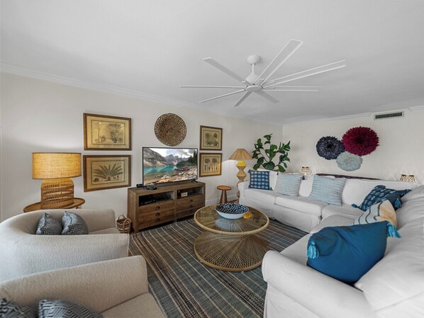Un salón con ventilador de techo blanco, televisor, sofá blanco, mesa de centro y varios objetos decorativos, incluyendo cuadros enmarcados y una planta.