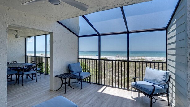 Un espacio al aire libre techado con vista a la playa, que incluye una mesa, sillas, un ventilador de techo y una barandilla.