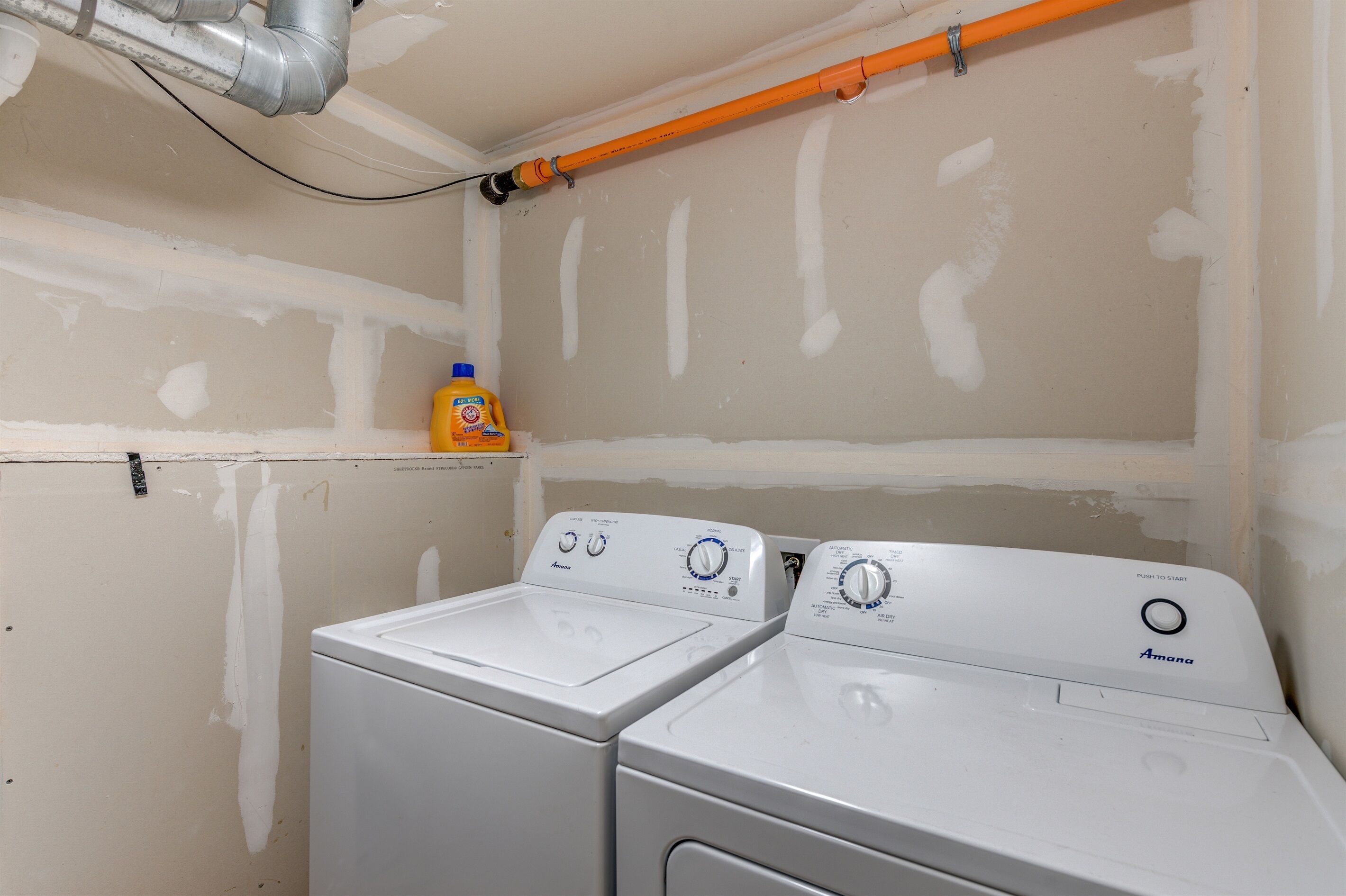 Une buanderie équipée de deux machines à laver Amana blanches, d’une étagère sur laquelle se trouve une bouteille de lessive, et dont les conduits apparents sont visibles.