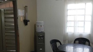 Una habitación con una mesa redonda y cuatro sillas, un dispensador de agua, una puerta con persiana metálica y una ventana con cortinas.