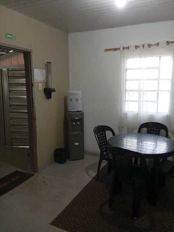 A room with a round table and four chairs, a water dispenser, a door with a metal shutter, and a window with curtains. - Pousada e Alojamento Recanto São José (Rio Verde)