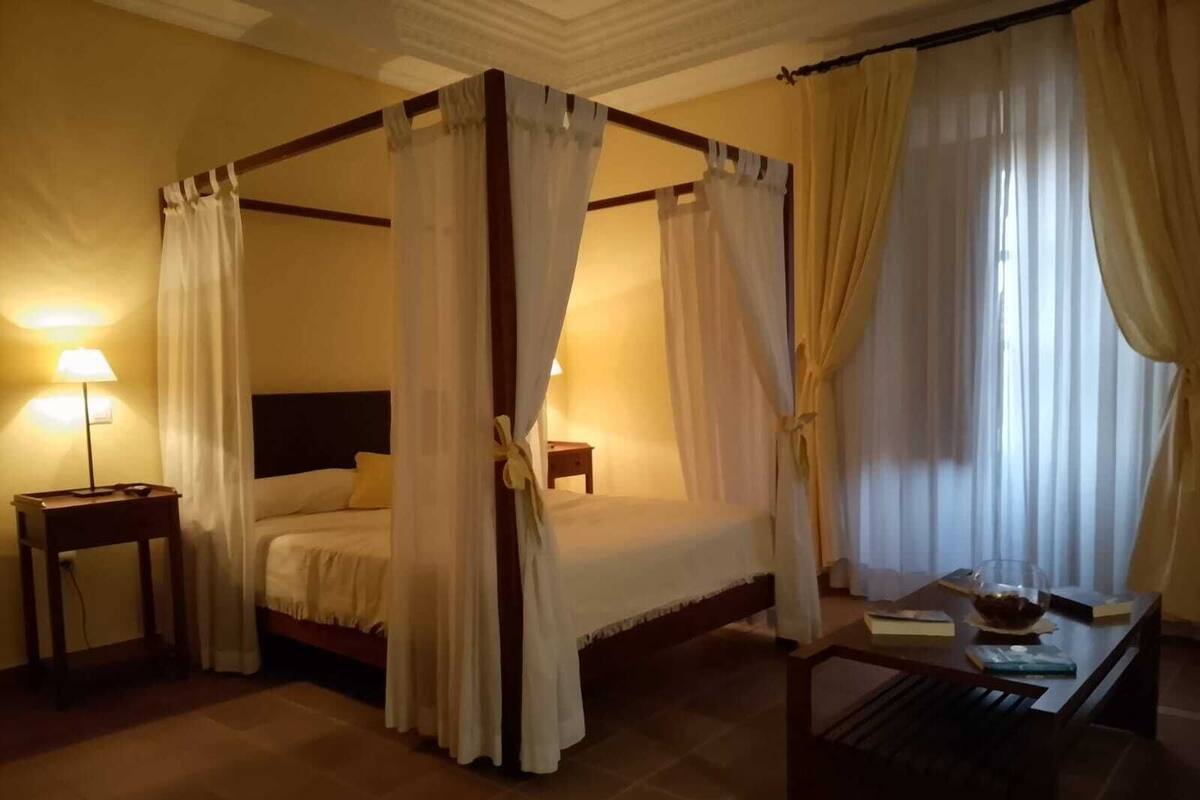 A four-poster bed with white linens, a bedside table with a lamp, and a small table with a bowl and books.