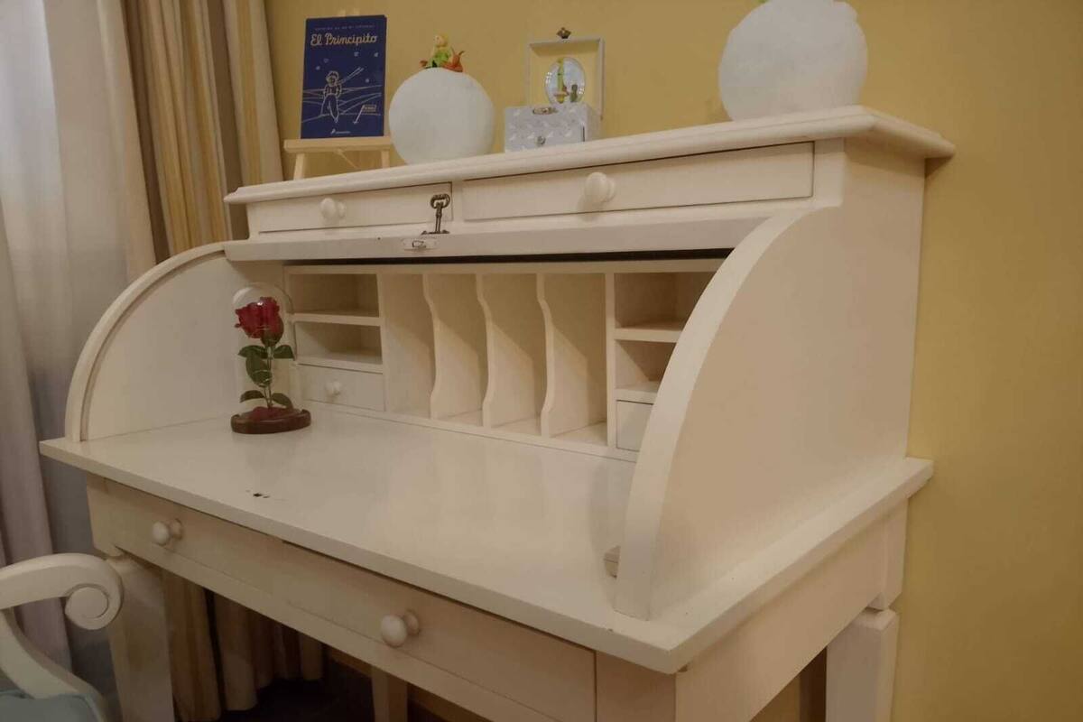 A white desk with a curved backrest, a drawer, and a shelf with decorative items.