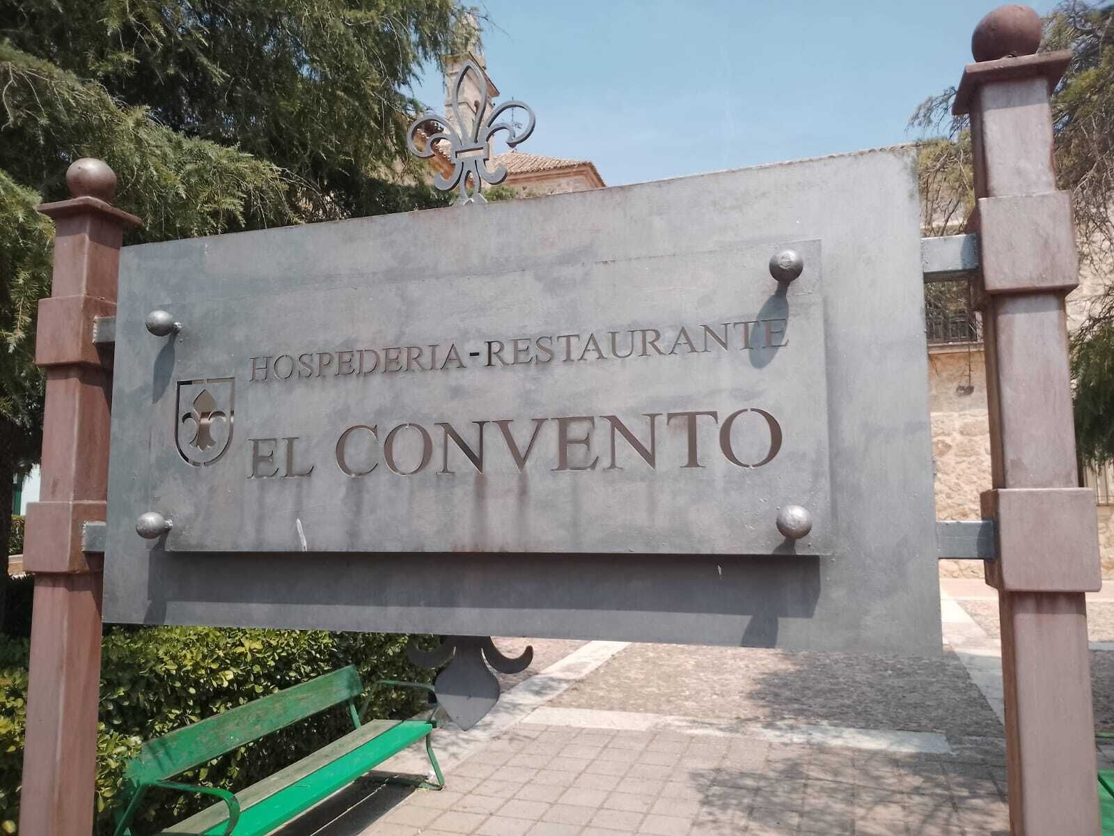 Señalización para 'HOSPEDERIA-RESTAURANTE EL CONVENTO', con un escudo de armas y una banca cerca.