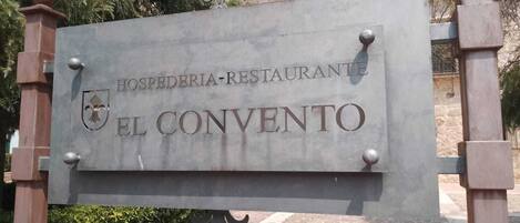 Opastekyltti, jossa lukee ”HOSPEDERIA-RESTAURANTE EL CONVENTO”, ja sen yhteydessä vaakuna sekä lähellä oleva penkki.