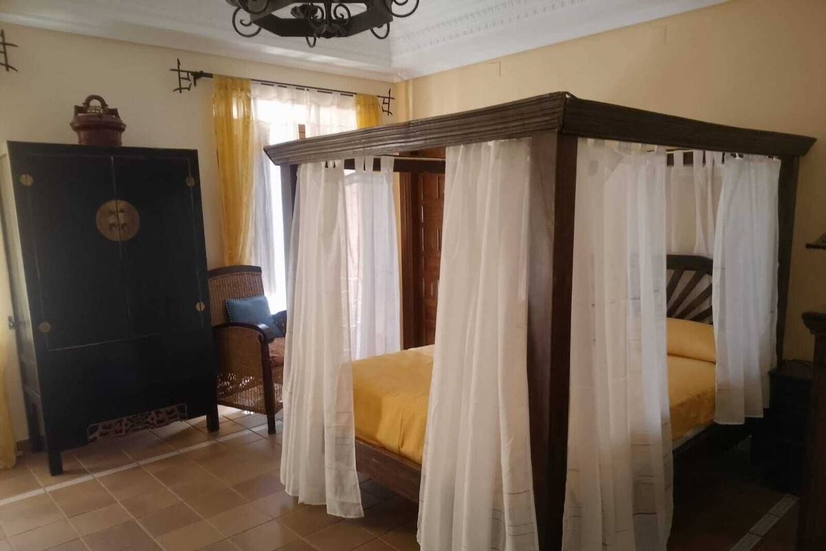 A traditional four-poster bed with white curtains, a wooden cabinet, and a wicker chair in a room with tiled flooring.