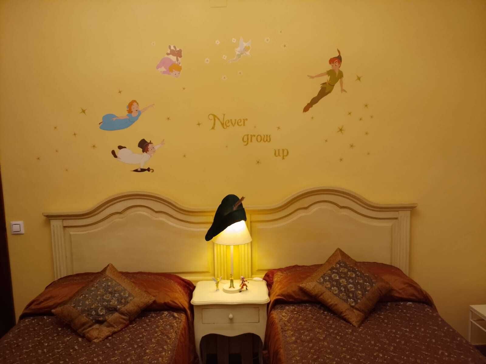 A hotel room with two beds, a nightstand with a lamp, and a wall decorated with fairy tale characters and the phrase "Never grow up".