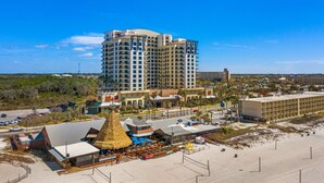 Un hotel de gran altura con varios pisos, una zona de playa con una estructura de techo de paja y un estacionamiento.