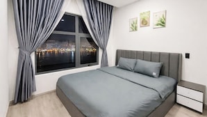 A modern bedroom with a large bed, grey curtains, a night view of a cityscape, and a nightstand. - Monarchy Luxury Serviced Apartments (Da Nang)