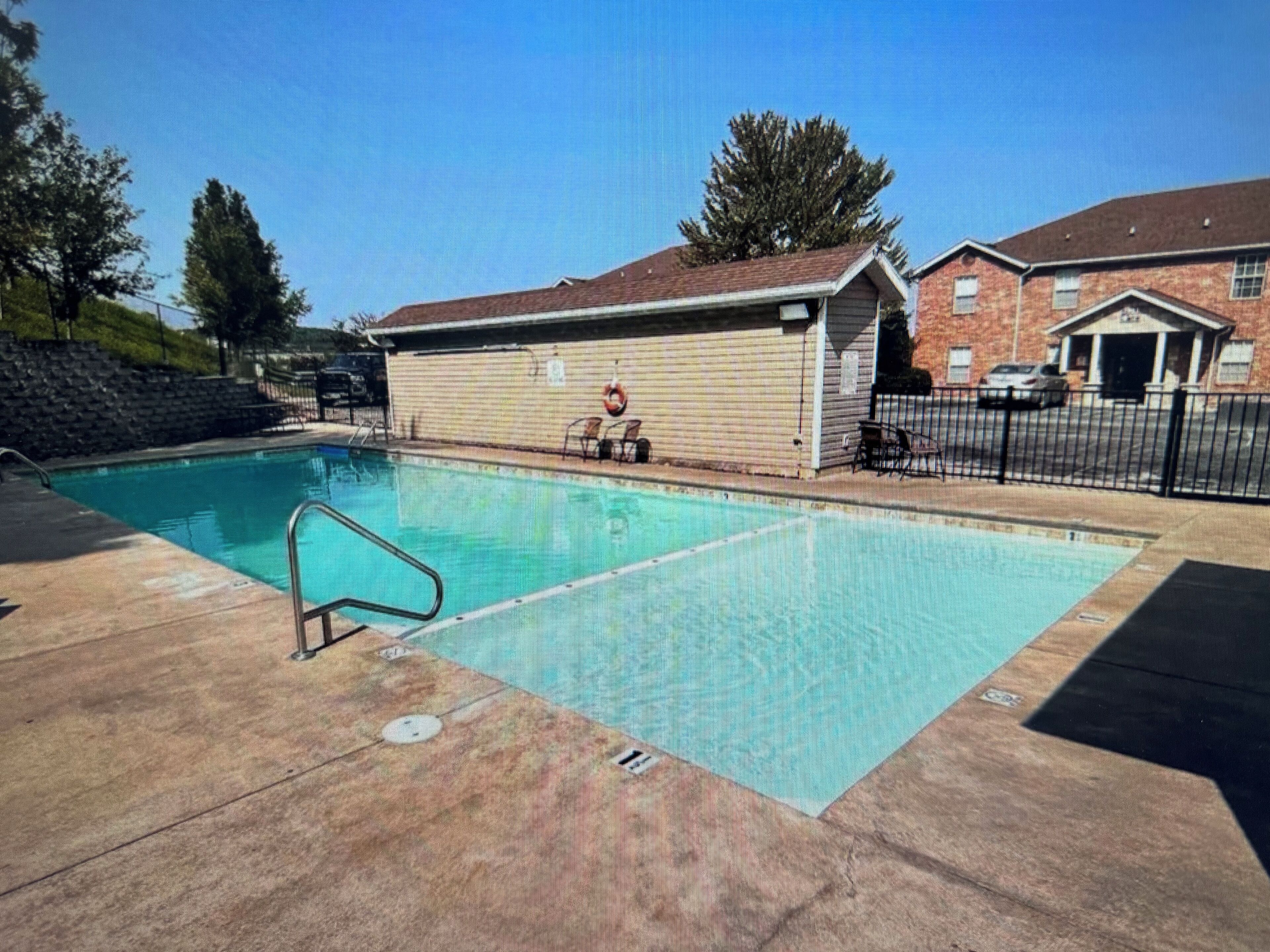 A swimming pool with a lifeguard chair and a lifebuoy, surrounded by a brick building and a fence.