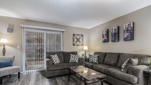 A living room with a grey sofa, a coffee table, and a white-framed window with blinds.