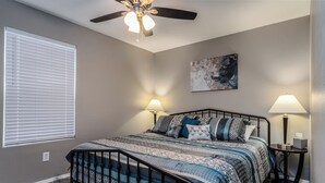 A bedroom with a bed, two bedside lamps, a ceiling fan, a painting on the wall, and a window with blinds.