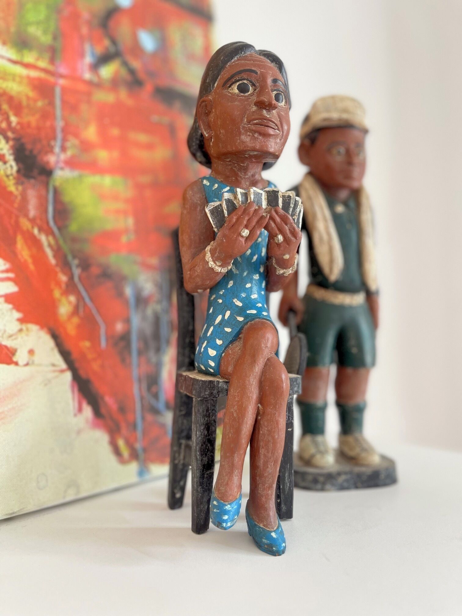 Two wooden figurines, one seated and one standing, with a colorful abstract painting in the background.