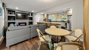Ein modernes Wohnzimmer mit einem grauen Ecksofa, einem runden Holzesstisch, Korbstühlen und einer Küchenzeile mit Kühlschrank und Schränken.