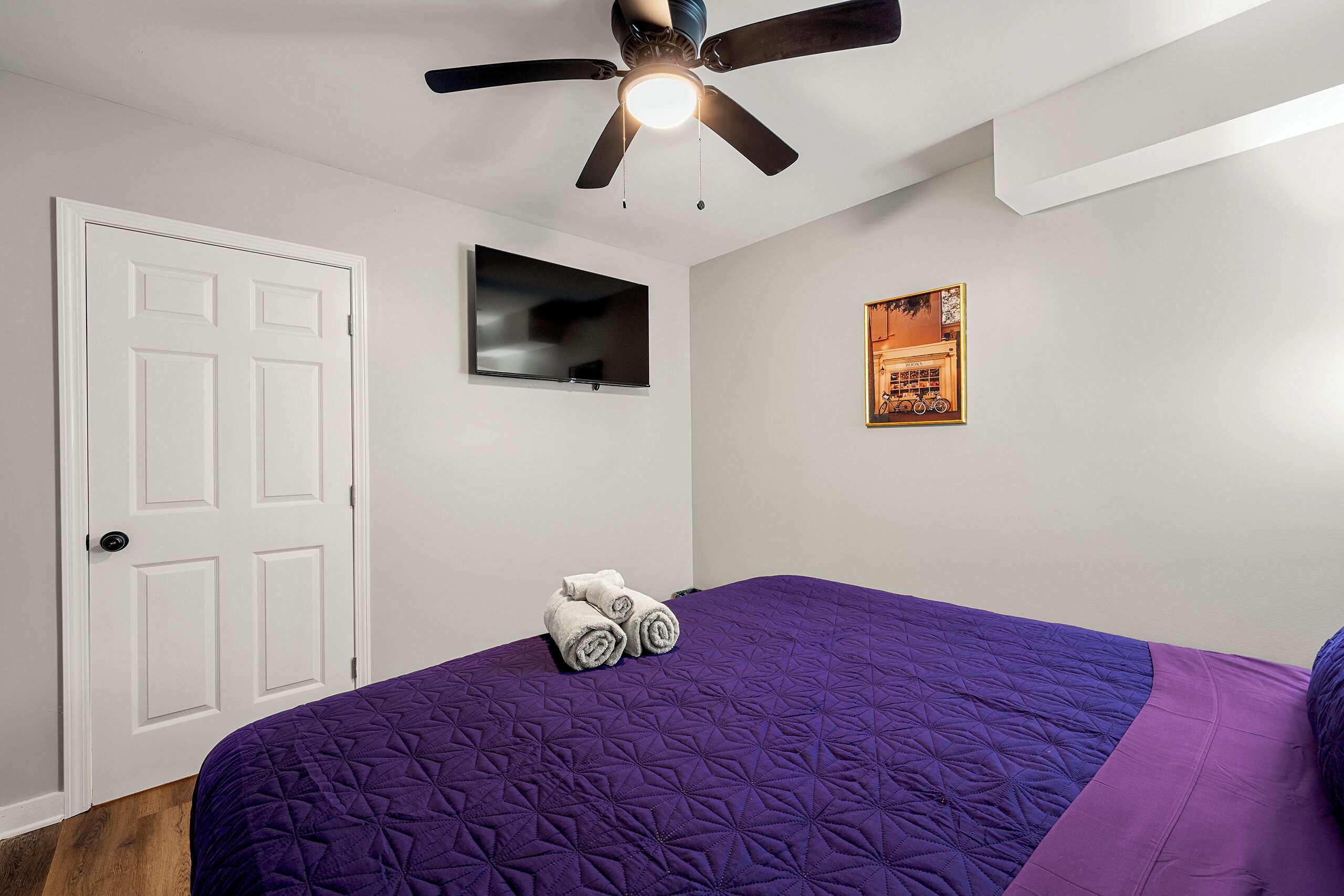 Ein Schlafzimmer mit einem violetten gesteppten Bettbezug, einem Deckenventilator, einem Flachbildfernseher, einer Tür und einem gerahmten Bild an der Wand.