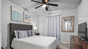 A bedroom with a tufted headboard, a bed with striped bedding, a ceiling fan, a television, a dresser, a nightstand with a lamp, and two framed artworks on the wall. - The VB Haven (Van Buren)