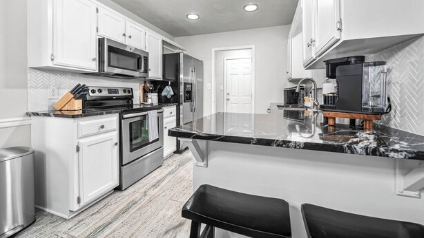 A modern kitchen with white cabinetry, stainless steel appliances, a marble countertop, and a built-in island with bar stools. - The VB Haven (Van Buren)