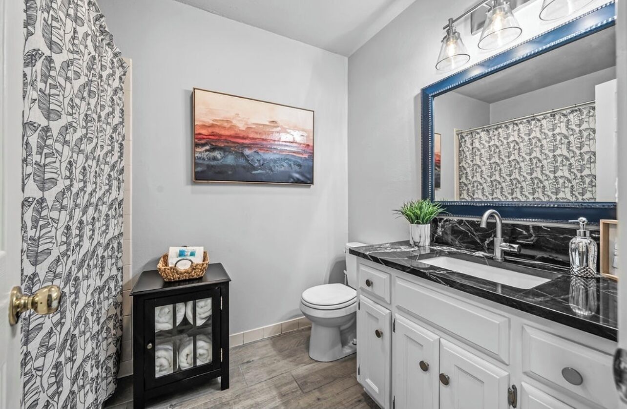 A bathroom with a black and white patterned shower curtain, a white toilet, a black cabinet with glass doors, a marble sink with a large mirror, and a framed painting on the wall.
