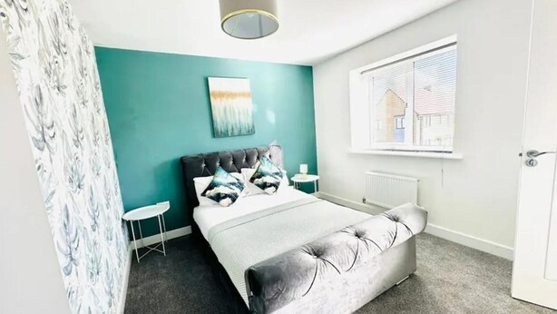 A bedroom with teal walls, a bed with a grey upholstered headboard, a bedside table, and a window with blinds.