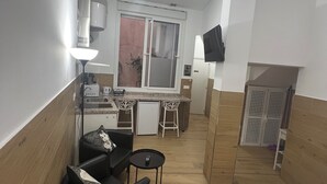 A compact hotel room with a bed, a small seating area with a table, a kitchenette, a TV mounted on the wall, and a sliding door leading to a bathroom.
