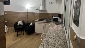 A compact kitchen with a built-in stove, microwave, and a stainless steel range hood. There is a small dining area with a chair and a table, and a window with a view of the outside.