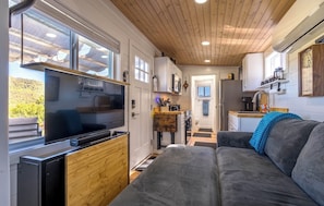 Cocina compacta con televisor, muebles de madera y ventana con vistas a la vegetación.