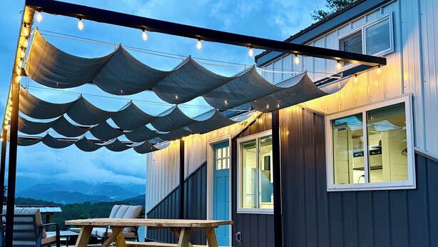Een houten terras met een jacuzzi, een picknicktafel en een overdekte zithoek met lichtjes.