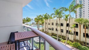 A balcony with a view of a swimming pool, palm trees, and a high-rise building.