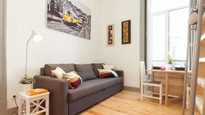 Una sala moderna con un sofá gris, una mesita auxiliar blanca con una planta, un escritorio con una silla y un ventanal con vistas a edificios.