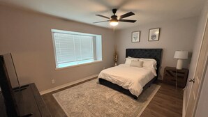 A bedroom with a bed, a ceiling fan, a window with blinds, a nightstand with a lamp, and a small wooden cabinet. - Updated Westbury Getaway Gem - 3 bedroom oasis (Houston)