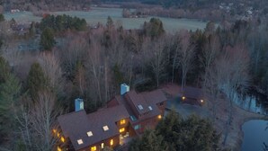 Una casa grande de varios pisos con una chimenea destacada, rodeada de un bosque denso y un lago.