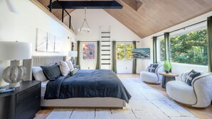 Un dormitorio moderno con una cama grande, un sofá, una mesita y vistas al exterior a través de amplios ventanales.