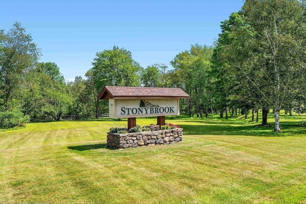 A sign for Stonybrook with a rustic wooden structure and stone base, surrounded by a well-maintained grassy area and trees.