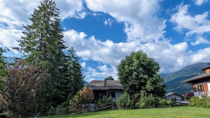 A traditional chalet with a sloped roof, surrounded by lush greenery and a clear blue sky.