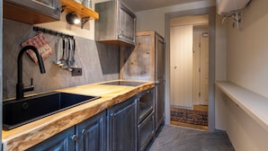 A narrow kitchen with dark wood cabinets, a black sink, and a wooden countertop. There is a hanging pot rack above the sink and a small shelf with jars.