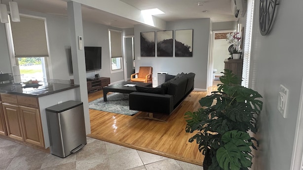 A modern living room with a black sofa, a coffee table, and a TV. There is a kitchen area with a refrigerator and a sink.