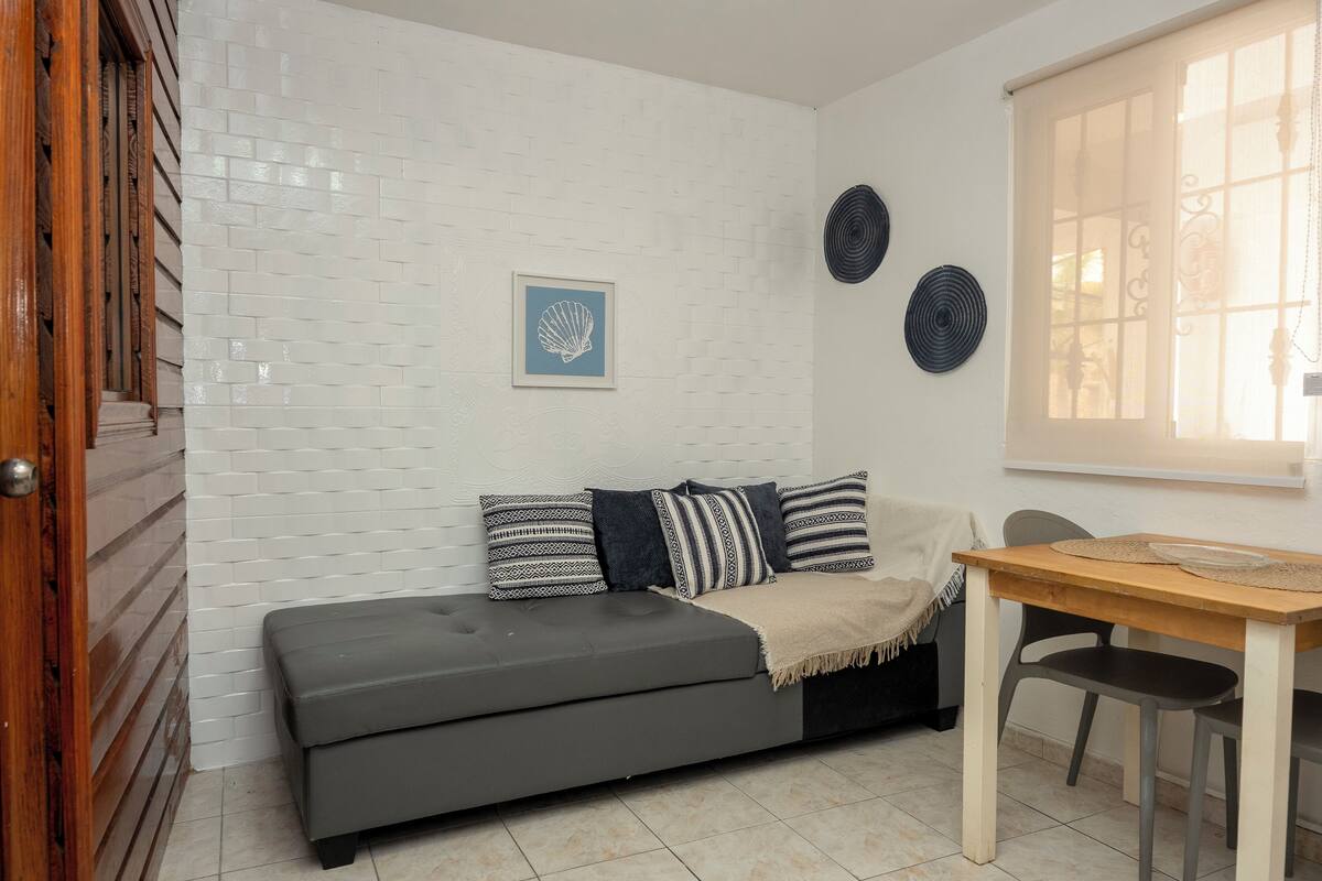 Een knus woongedeelte met een grijze bank, een houten eettafel en een witte bakstenen muur.