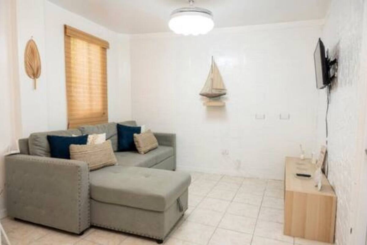 Een woonkamer met een grijze hoekbank, een klein houten bijzettafeltje en een aan de muur bevestigde televisie.