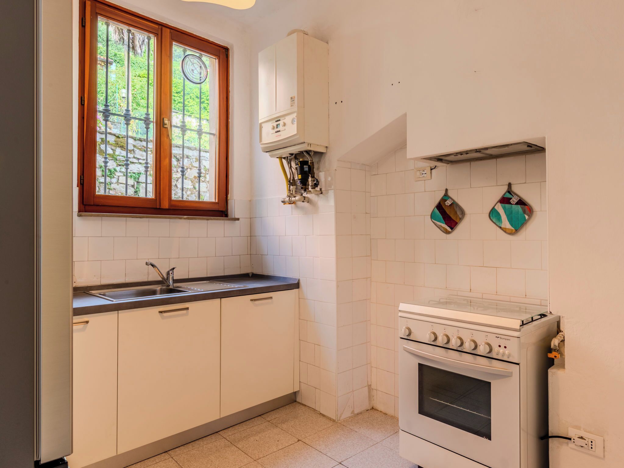 Eine Küche mit weißen Schränken, einem Fenster mit Blick auf Grünflächen, einem Herd und einem farbenfrohen Geschirrtuch an der Wand.