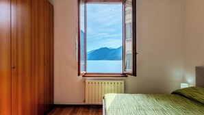 A bedroom with a wooden wardrobe, a radiator, a bed with a striped bedspread, and a window with a view of a lake and mountains. - Martino by Interhome (Argegno)