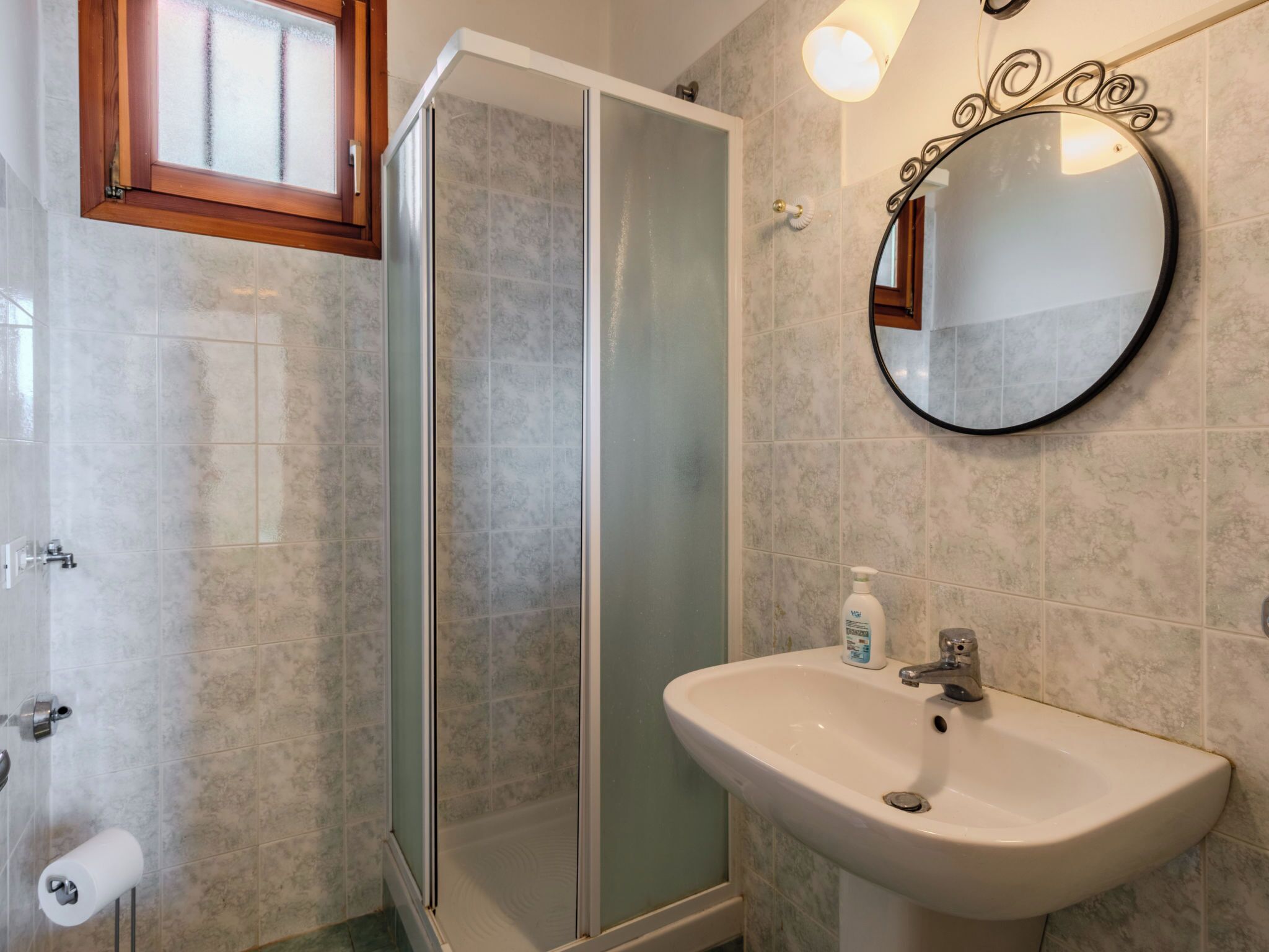 A bathroom with a glass shower enclosure, a white sink, and a round mirror with a decorative frame.