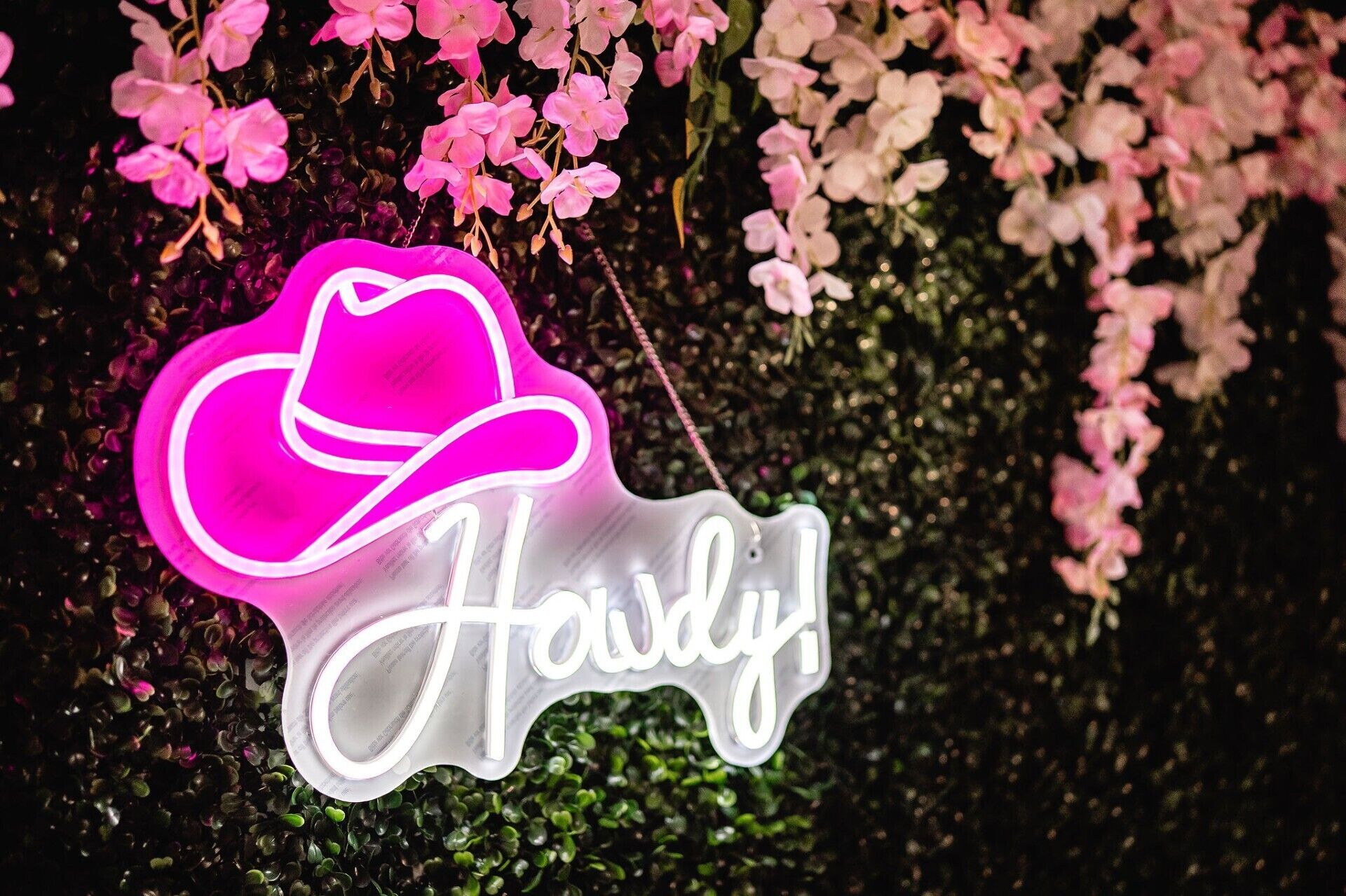 Um letreiro de neon com um chapéu de cowboy rosa e a palavra 'Haidy!', com um fundo de flores e folhagem rosa.