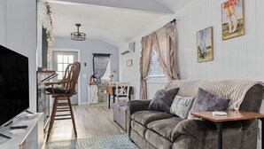 A living room with a sofa, a TV, a wooden chair, a dining table with chairs, and a kitchen area in the background. - Tiny House with lake view and large yard (McMinnville)
