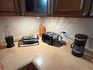 Uma bancada de cozinha com liquidificador, torradeira, cafeteira e um porta-utensílios de madeira.